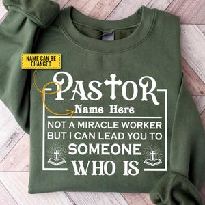 May include: An olive green sweatshirt with the word "PASTOR" in large white letters. Below, the text reads "Name Here", "NOT A MIRACLE WORKER BUT I CAN LEAD YOU TO SOMEONE WHO IS". A yellow box says "NAME CAN BE CHANGED".