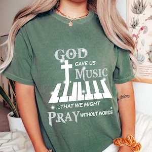Peut inclure: T-shirt vert avec un motif blanc qui dit "Dieu nous a donné la musique... pour que nous puissions prier sans paroles". Le motif comprend une croix et un clavier de piano.
