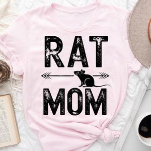 a pink t-shirt with the text "RAT MOM" printed on it, along with a graphic of a rat. The t-shirt is placed on a bed, and there are various items such as a hat, a book, and a cup nearby.