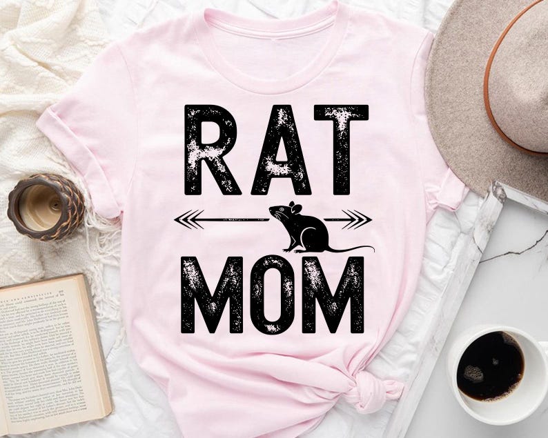 a pink t-shirt with the text "RAT MOM" printed on it, along with a graphic of a rat. The t-shirt is placed on a bed, and there are various items such as a hat, a book, and a cup nearby.