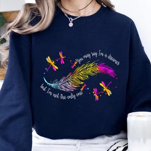 May include: Navy blue sweatshirt featuring a colorful feather and dragonfly design. The text reads "You may say I'm a dreamer but I'm not the only one." The design includes pink, yellow, and purple accents.