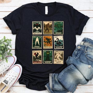 May include: Black t-shirt featuring a collection of vintage-style postage stamps with cryptid illustrations. The stamps depict creatures like Mothman, Bigfoot, and the Loch Ness Monster. The shirt is paired with blue jeans and white sneakers.