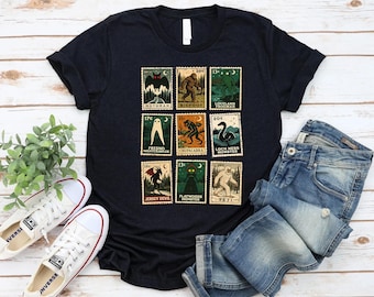 May include: Black t-shirt featuring a collection of vintage-style postage stamps with cryptid illustrations. The stamps depict creatures like Mothman, Bigfoot, and the Loch Ness Monster. The shirt is paired with blue jeans and white sneakers.