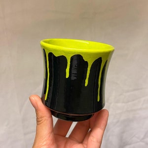 May include: A black ceramic cup with a neon green interior and dripping paint design. The cup is held in a hand and has a rounded shape. The cup is approximately 3.5 inches tall.