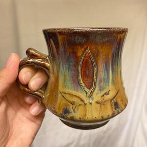 Puede incluir: Una taza de cerámica marrón y amarilla con un esmalte único y un diseño de hojas. La taza tiene un asa y una forma redondeada.