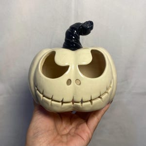 Puede incluir: Un recipiente de cerámica en forma de calabaza de color crema con un tallo negro. La calabaza tiene grandes recortes para los ojos y la boca, y dos pequeños orificios nasales circulares. El recipiente está diseñado con un tema de Halloween.