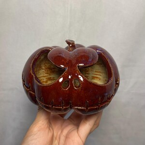 Puede incluir: Un recipiente de cerámica con forma de calabaza con un esmalte rojo oscuro brillante. La calabaza tiene ojos, boca y tallo tallados. El interior es de un color beige claro. El recipiente está diseñado para la decoración de Halloween.
