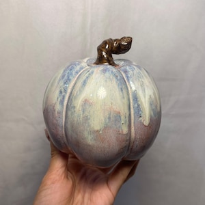 Peut inclure: Une citrouille en céramique avec une finition brillante. La citrouille est principalement bleue et blanche, avec des touches de rouge et de marron. La tige est de couleur marron foncé. La citrouille a un design côtelé et est un objet décoratif.