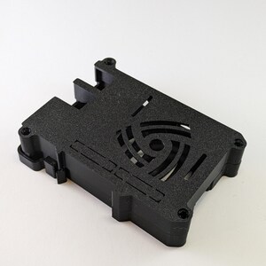 Könnte beinhalten: Schwarzes Plastikgehäuse mit einem Lüfterausschnitt und einem kreisförmigen Design in der Mitte. Das Gehäuse ist für einen Raspberry Pi konzipiert.