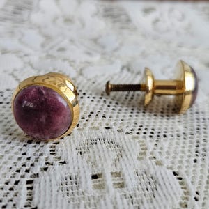 May include: Two gold-toned cabinet knobs with a round, purplish-red stone center. One knob is fully assembled, while the other is disassembled, showing the screw and backplate. The knobs are set against a white lace background.