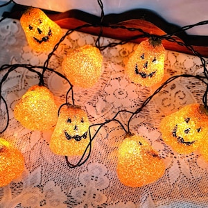 May include: A string of orange pumpkin-shaped lights connected by a black wire. Each pumpkin features a textured, glittery surface with a carved jack-o'-lantern face, including eyes and a mouth, illuminated from within by a warm glowing light.