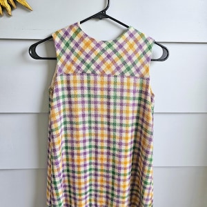 May include: A sleeveless dress with a yellow, green, and purple plaid pattern. The dress is hanging on a black hanger. The dress has a scoop neckline. The background is a white wall.