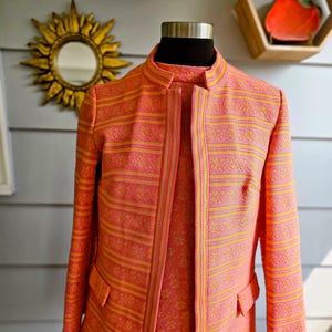 May include: A pink and yellow patterned jacket with a mandarin collar and two front pockets. The jacket is displayed on a mannequin and features a striped design with floral accents. The lining is a lighter shade of pink with a floral pattern.