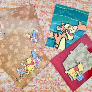 Puede incluir: Cuatro carpetas coloridas con el tema de Winnie the Pooh de Disney. Dos son de color marrón claro con patrones de hojas y pegatinas de personajes. Una es turquesa con Tigger, y otra es roja con Pooh sosteniendo una pelota.