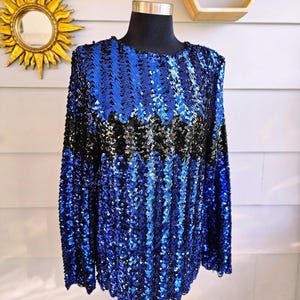 May include: A long-sleeved top covered in blue and black sequins. The sequins create a vertical striped pattern. The top is displayed on a black mannequin head and torso. The background includes a gold sunburst mirror and a wooden geometric shelf.