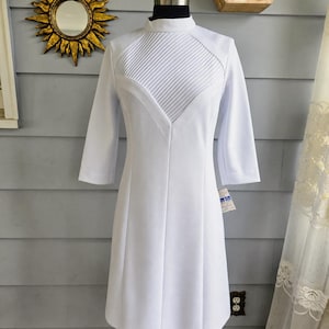 Pode incluir: Um vestido branco com gola alta e mangas três-quartos. O corpete apresenta um design plissado, e o vestido tem uma cintura ajustada e uma saia rodada. O vestido é exibido em um manequim, mostrando sua silhueta.