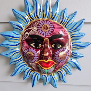 May include: A ceramic sun wall plaque featuring a deep red face with black eyes, arched eyebrows, and bright red lips. The face is decorated with purple and gold floral patterns. It has blue, yellow, and light blue rays and a white hanging cord.