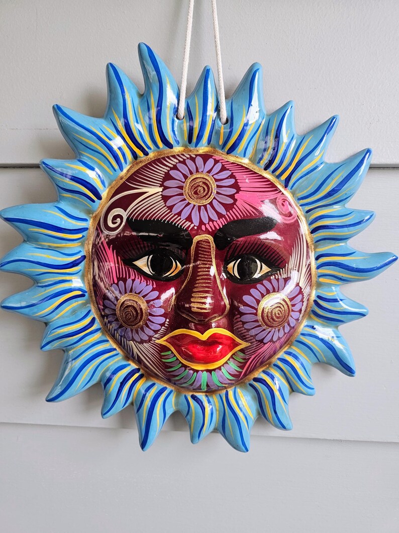 May include: A ceramic sun wall plaque featuring a deep red face with black eyes, arched eyebrows, and bright red lips. The face is decorated with purple and gold floral patterns. It has blue, yellow, and light blue rays and a white hanging cord.