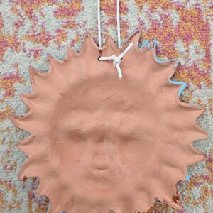 May include: A terracotta-colored ceramic sun wall plaque featuring a sculpted face with closed eyes and a mouth in a neutral expression. The sun has numerous pointed rays extending outward and includes a white cord loop for hanging purposes.
