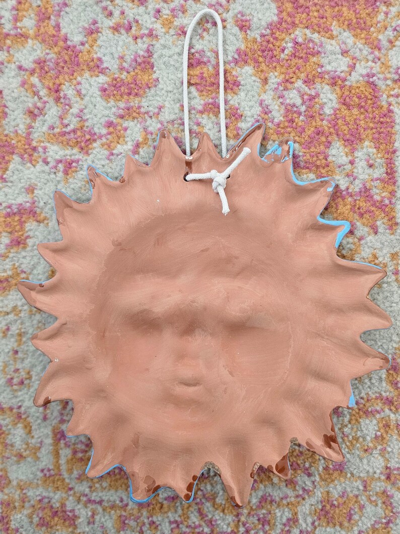 May include: A terracotta-colored ceramic sun wall plaque featuring a sculpted face with closed eyes and a mouth in a neutral expression. The sun has numerous pointed rays extending outward and includes a white cord loop for hanging purposes.