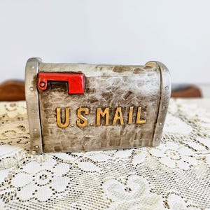 May include: A decorative mailbox with a distressed silver finish and a red flag. The words "U.S. MAIL" are in gold lettering on the front. The mailbox is sitting on a white lace tablecloth.