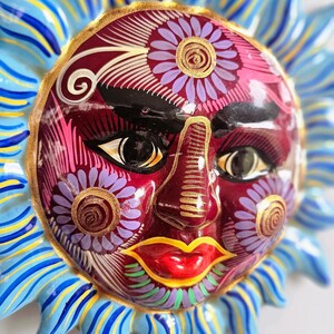 May include: A ceramic sun wall plaque featuring a deep red face with hand-painted floral details in purple, gold, and white. The sun has bright red lips, dark eyebrows, and stylized eyes. It is surrounded by radiating blue and yellow pointed rays.