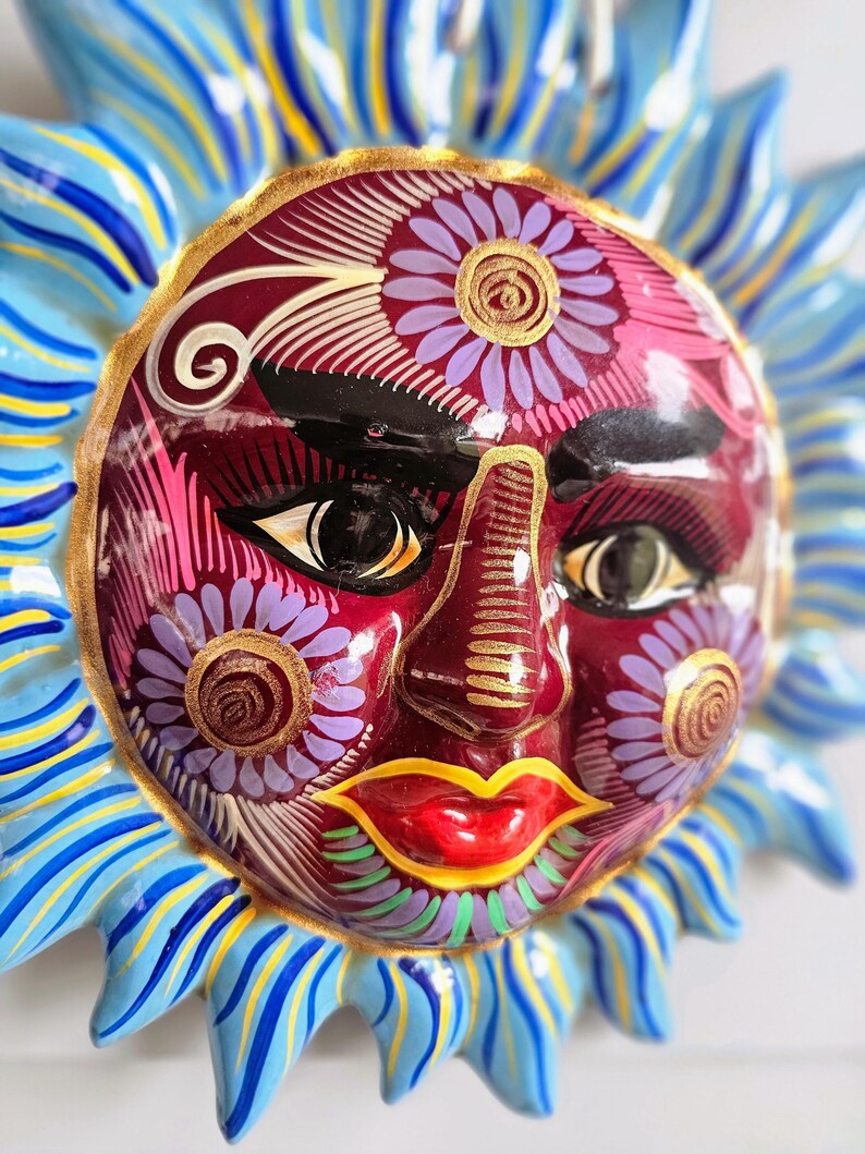 May include: A ceramic sun wall plaque featuring a deep red face with hand-painted floral details in purple, gold, and white. The sun has bright red lips, dark eyebrows, and stylized eyes. It is surrounded by radiating blue and yellow pointed rays.