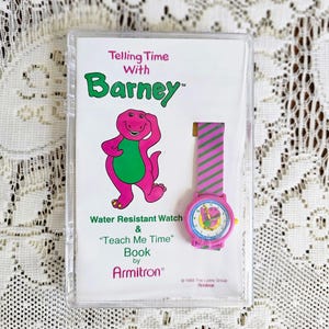 Può includere: Un set vintage di orologio e libro di Barney il Dinosauro. L'orologio è rosa con un cinturino a righe e un quadrante colorato. La copertina del libro presenta Barney e il testo "Telling Time With Barney".