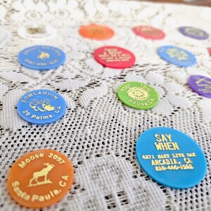 May include: A collection of small, round, colorful plastic tokens featuring various embossed gold text and graphics, including names of locations like Arcadia, CA, 29 Palms, CA, and Santa Paula, CA, along with specific organization names and phone numbers.