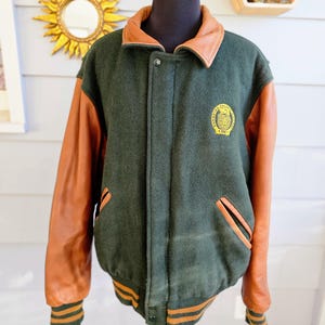 May include: Green and brown varsity jacket with leather sleeves. The jacket has a brown leather collar and a patch on the chest. The cuffs and waistband have orange stripes. The jacket is displayed on a mannequin.