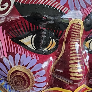 May include: A glossy, deep red decorative mask featuring intricate hand-painted details. The design includes black almond-shaped eyes with white accents, gold-painted nose ridges, and stylized floral patterns in lavender, gold, and white.