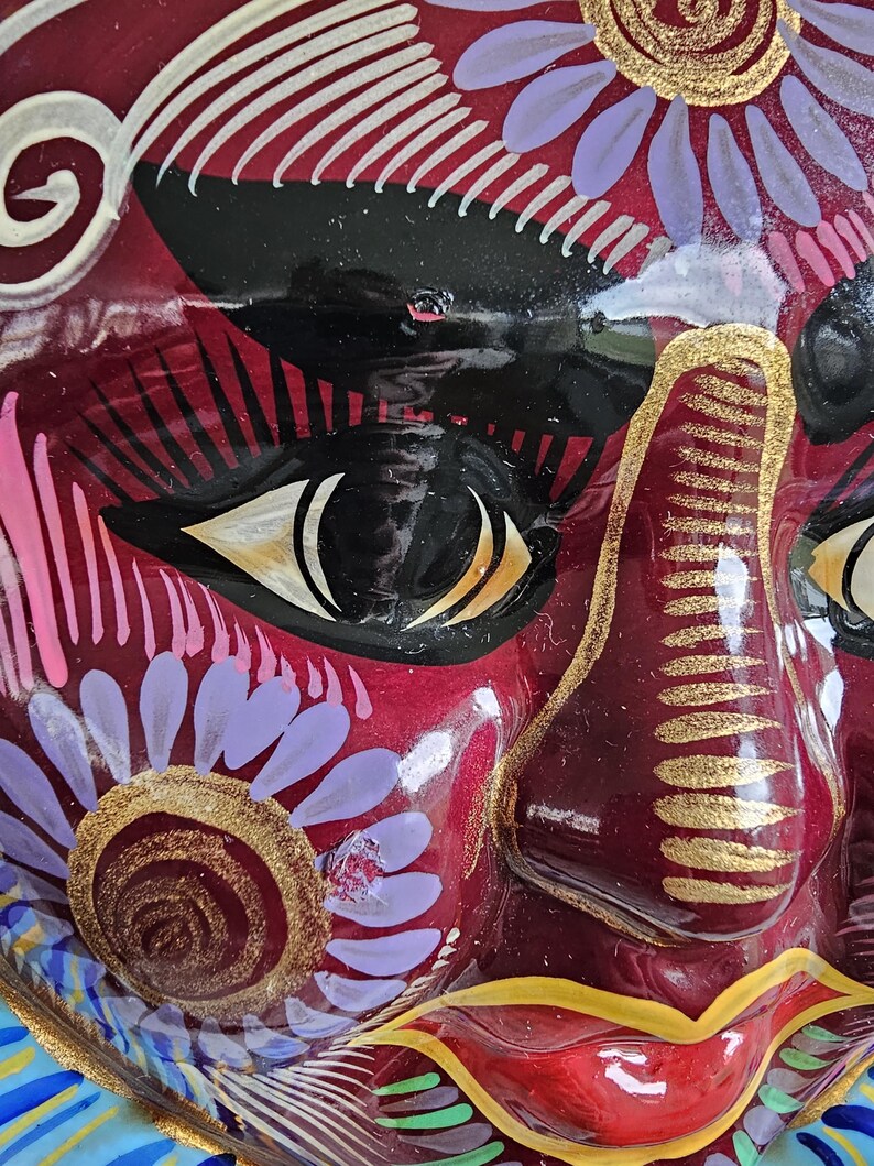 May include: A glossy, deep red decorative mask featuring intricate hand-painted details. The design includes black almond-shaped eyes with white accents, gold-painted nose ridges, and stylized floral patterns in lavender, gold, and white.