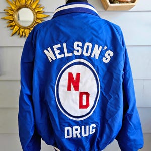 May include: A royal blue jacket with white and blue striped trim. The back of the jacket features the text "NELSON'S DRUG" in white lettering, with a circular logo containing the letters "N" and "D" in red.