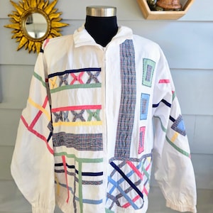 May include: White zip-up jacket with colorful geometric patchwork. The jacket features a stand-up collar, long sleeves, and a full zipper. The patchwork includes pink, blue, green, and yellow accents. A vintage style jacket.