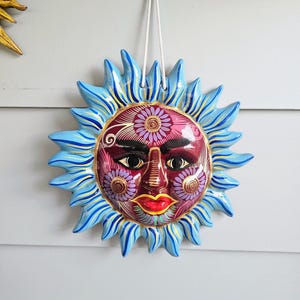 May include: A ceramic sun wall hanging featuring a deep red face with intricate purple and gold floral patterns, black eyes, and bright red lips. The sun rays are painted in shades of light blue with yellow accents, and a white cord is attached for hanging.