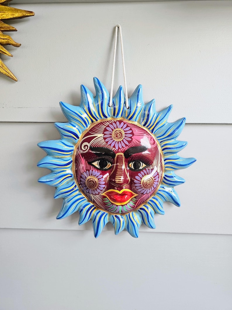 May include: A ceramic sun wall hanging featuring a deep red face with intricate purple and gold floral patterns, black eyes, and bright red lips. The sun rays are painted in shades of light blue with yellow accents, and a white cord is attached for hanging.