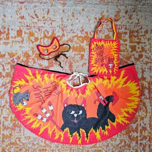 Puede incluir: Un conjunto de disfraz de diablo en rojo y naranja que incluye una capa, una máscara y una bolsa de tela. La capa muestra un gato negro con cuernos, un tridente y el texto 'A LITTLE HOT STUFF'. La bolsa presenta un pequeño diablo rojo y el mismo texto.