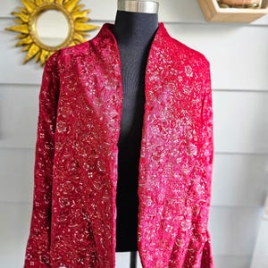 May include: A red, long-sleeved jacket with a floral pattern. The jacket is displayed on a black mannequin. The fabric has a textured appearance with gold accents. The jacket has a shawl collar and a slightly flared sleeve.