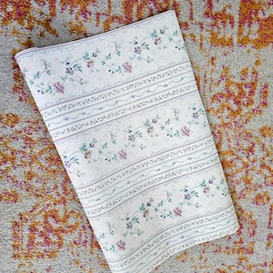 May include: A roll of off-white wallpaper featuring a repeating pattern of delicate floral vines with pink and purple blossoms, interspersed with thin horizontal stripes and wavy lines in muted tones across the entire surface of the paper.