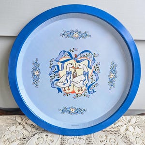 Puede incluir: Una bandeja redonda de color azul claro con un borde azul brillante. La bandeja presenta un diseño central de dos gansos blancos con lazos azules, rodeados de motivos florales y cintas. Un objeto decorativo vintage.