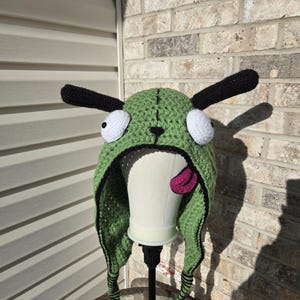 May include: Green crocheted hat with black ears and white eyes. The hat has a pink tongue sticking out and black and green stripes on the ties.