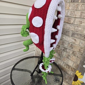May include: A crocheted red and white plant monster hat with green vines. The hat has a large mouth with white teeth and a red tongue.