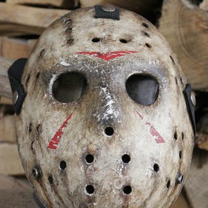 Może przedstawiać: Biała maska hokejowa z czerwonymi oznaczeniami i wieloma otworami. Maska jest zużyta i ma postarzony wygląd.