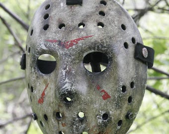 Könnte beinhalten: Eine verwitterte Eishockeymaske mit abgenutzter grauer Oberfläche, dunklen Flecken und roten Chevron-Markierungen. Die Maske verfügt über mehrere kreisförmige Belüftungslöcher, zwei Augenöffnungen und schwarze elastische Bänder, die seitlich mit Metallnieten befestigt sind.