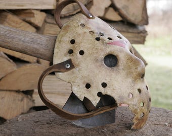 May include: A white plastic hockey mask with a red stripe and holes throughout. The mask is sitting on a wooden surface with a wooden axe handle in the background.