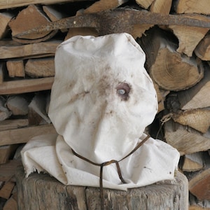 May include: A white fabric mask with a single eye opening and brown stains, tied with a brown leather cord. The mask is placed on a wooden stump, with a backdrop of stacked firewood.