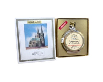 Puede incluir: Una botella redonda de vidrio transparente de colonia Original-Haus con tapón dorado, presentada en una caja dorada. La etiqueta presenta un emblema decorativo y texto. Una tarjeta con "Russian Leather" y una imagen de catedral está al lado.
