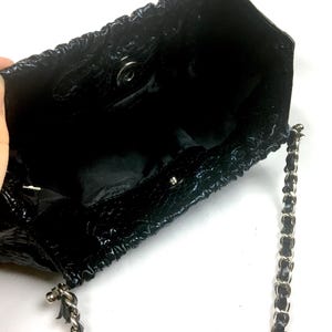 Puede incluir: Un bolso de mano de charol negro con una correa de cadena. El bolso tiene un dise&ntilde;o fruncido y un cierre plateado.