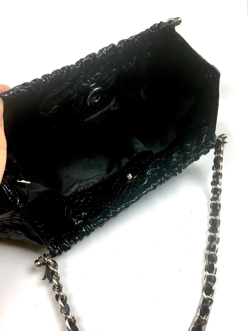 Puede incluir: Un bolso de mano de charol negro con una correa de cadena. El bolso tiene un dise&ntilde;o fruncido y un cierre plateado.