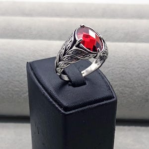 Op de afbeelding: Een zilveren ring met een grote, facetgeslepen, rode edelsteen. De ring heeft een sierlijk ontwerp met bladeren op de band. De edelsteen is gezet in een klauwzetting.